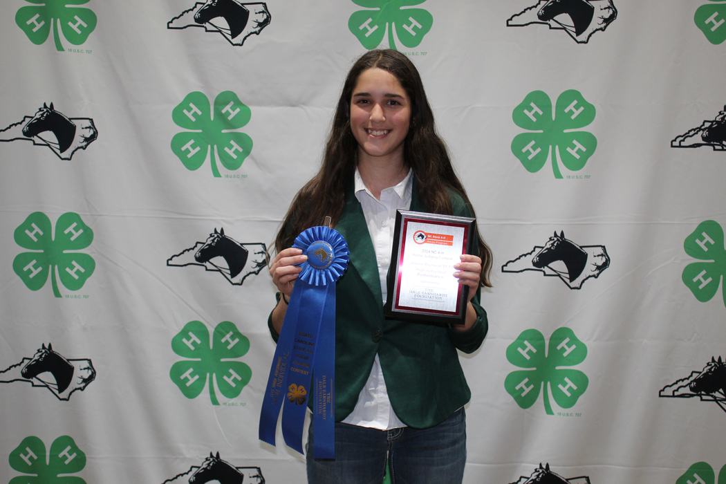 Young person holding blue ribbon and plaque in front of 4-H clover backdrop