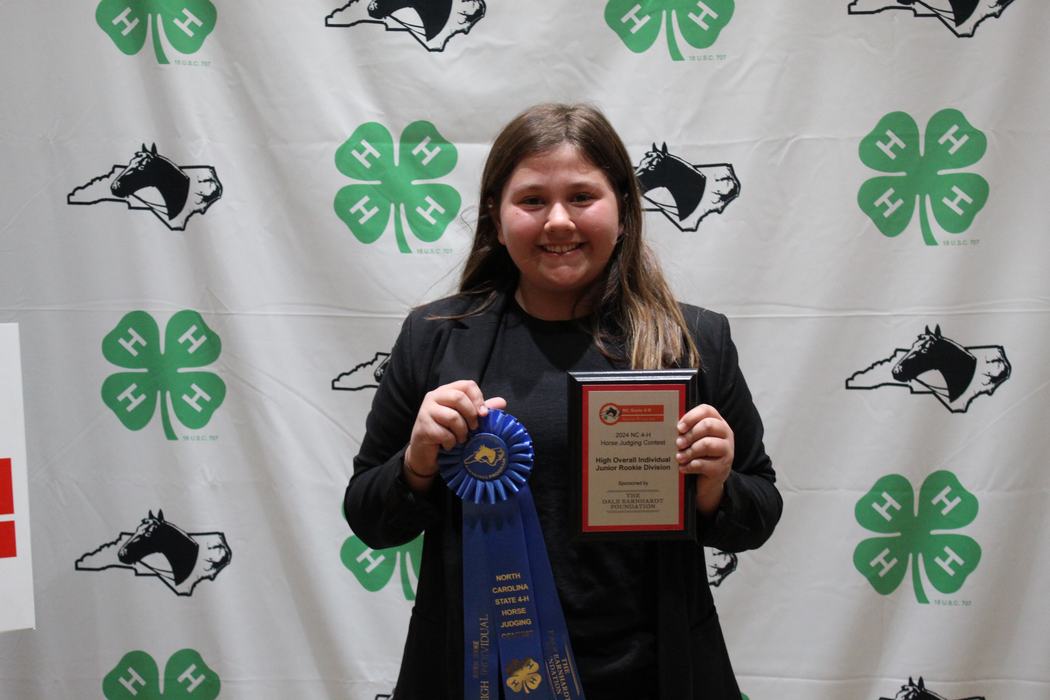 Young person holding blue award ribbon and plaque in front of 4‑H clover backdrop