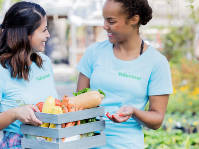 Two volunteers exchanging a crate of vegetables; shirts read "Volunteer" and banner reads "Using SNAP at the Farmers' Market"