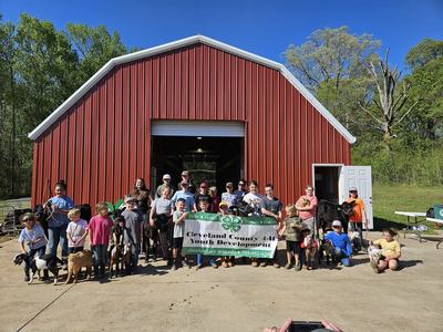 Youth with goats and cows in front of red barn, banner "Cleveland County 4‑H Youth Development"