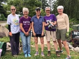 Five women standing outdoors by garden rocks; two wear purple shirts reading "WALK."