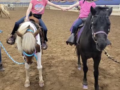 Two children wearing helmets holding hands while sitting on ponies in an indoor arena