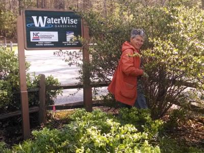 Person in orange jacket pruning shrubs beside a "WaterWise Gardening" sign