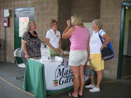 Five women at a table with a "Master Gardeners" banner and a "Garden Questions" sign.