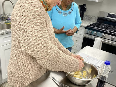 Two women in a kitchen mixing sliced bananas in a metal bowl on a stainless counter.