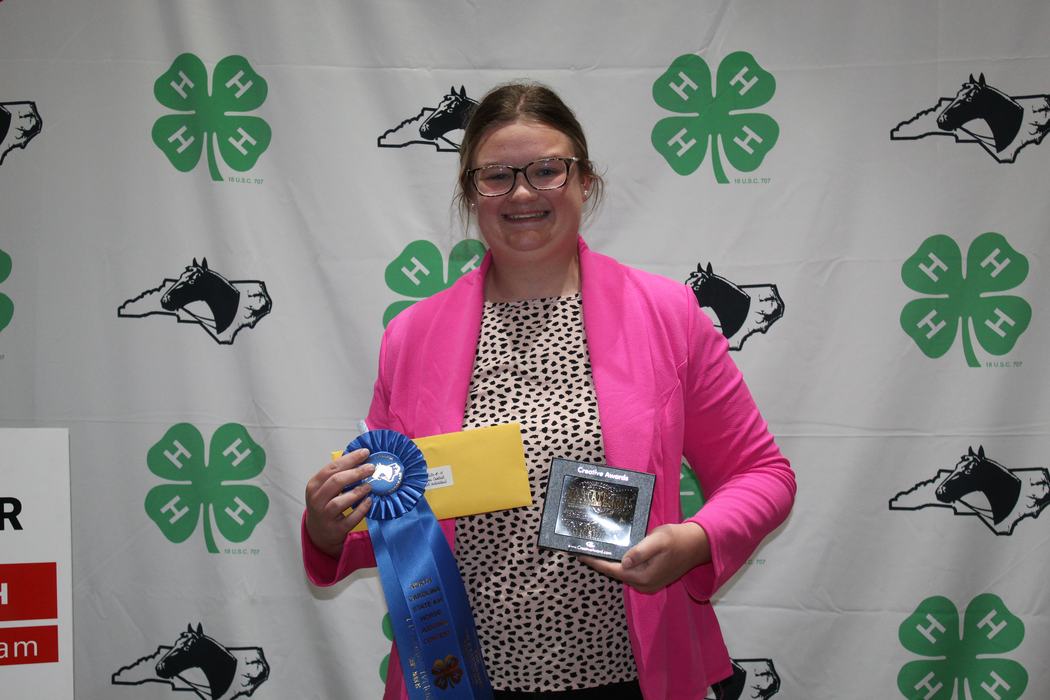 Young person holding blue ribbon, yellow envelope, and plaque in front of 4‑H clover backdrop