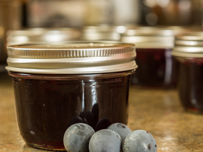 Jar of dark jam on countertop with four dusty blue grapes in front
