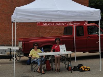 Two people seated at a table under a canopy with banner "Master Gardener Volunteers" and a red pickup behind