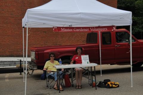 Two people seated at a table under a canopy with banner "Master Gardener Volunteers" and a red pickup behind