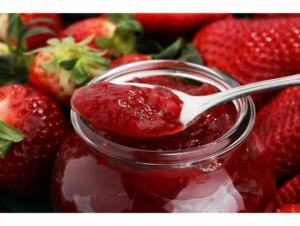 Jar of strawberry jam with a spoonful above the jar and whole strawberries around