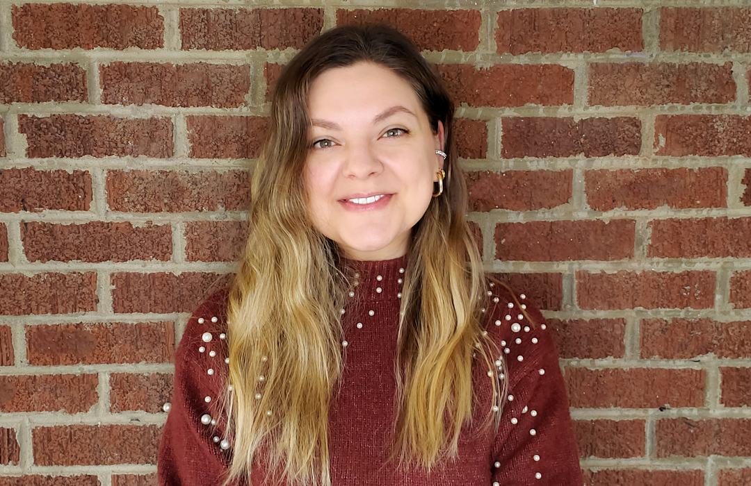 Person with long blond hair in maroon sweater decorated with pearls, standing against brick wall