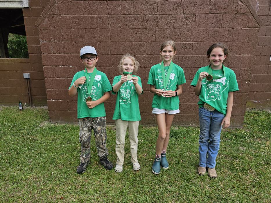 Four children in green shirts showing medals while standing against a brown cinderblock wall