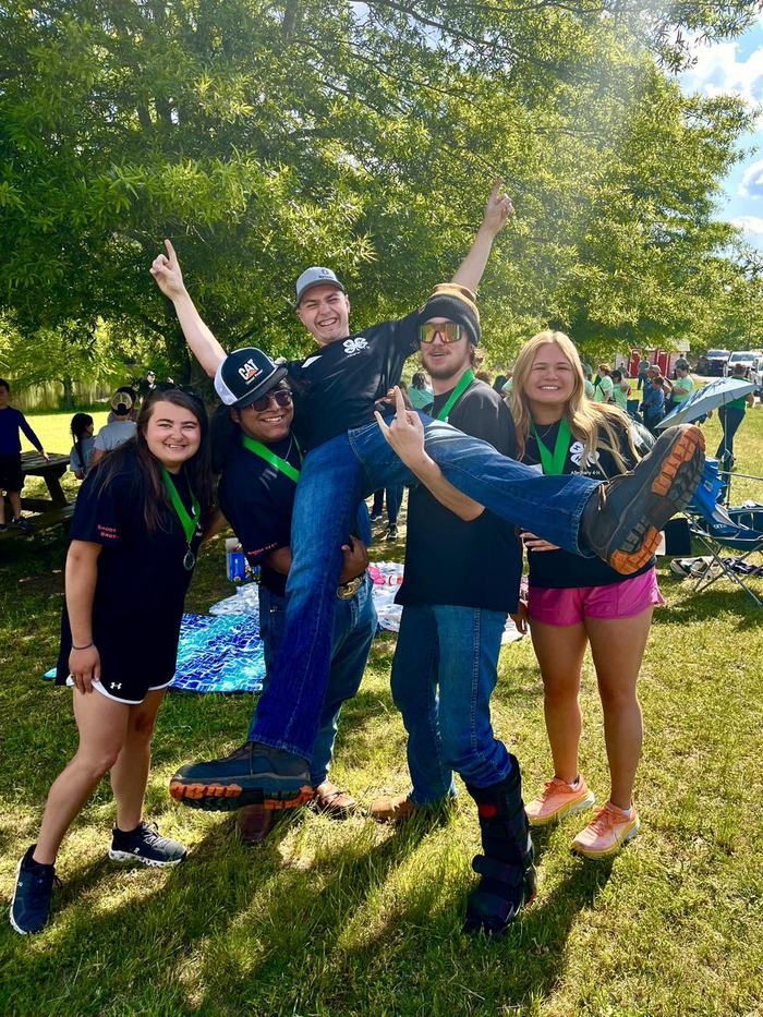 Five teens wearing medals, three holding a fourth teen horizontally in a park