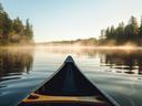 Canoe bow on calm misty lake at sunrise with tree-lined shore reflections