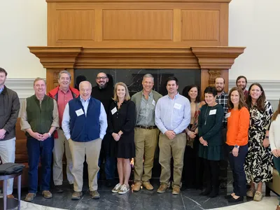 About 15 adults standing indoors in front of a large wooden mantel and fireplace.