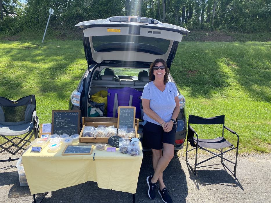 Woman standing beside open car trunk selling cookies at table with Creations Cookie Co sign