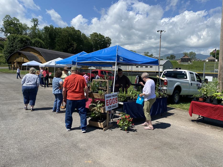 Outdoor farmers market tents with shoppers; sign reading "FARM FRESH EGGS FOR SALE."