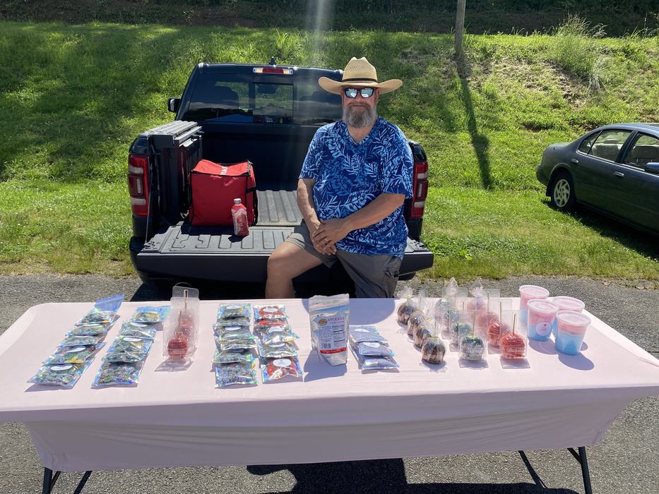 Man wearing straw hat and sunglasses on pickup tailgate behind table of packaged candies