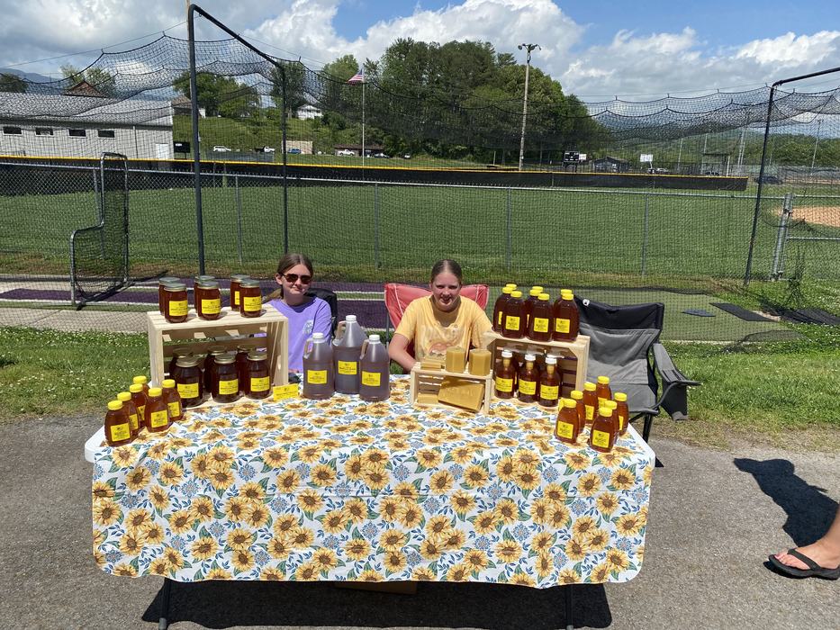 Two young vendors selling jars and bottles of honey at an outdoor table with sunflower cloth