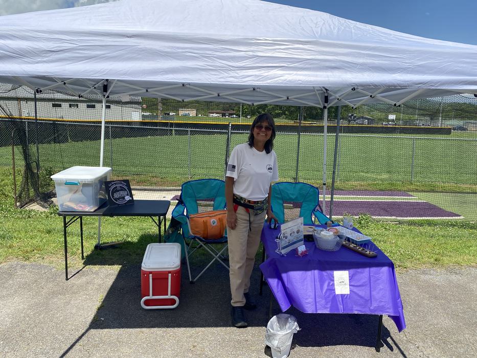 Woman standing at a canopy booth with tables, chairs and supplies beside a baseball field