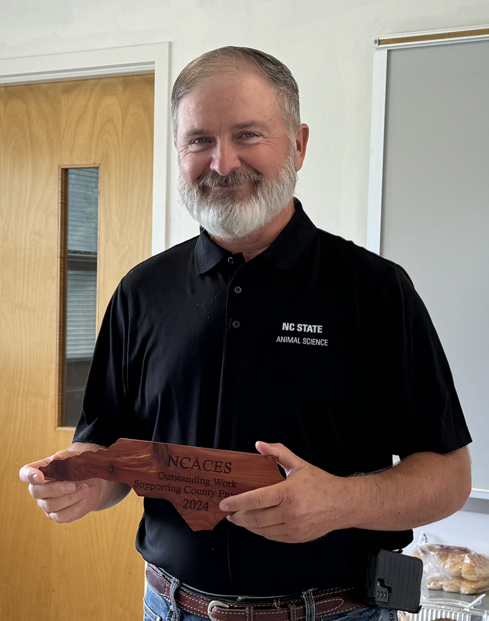Gentleman in a black polo with NC State logo holds an award made of wood and shaped like North Carolina. 