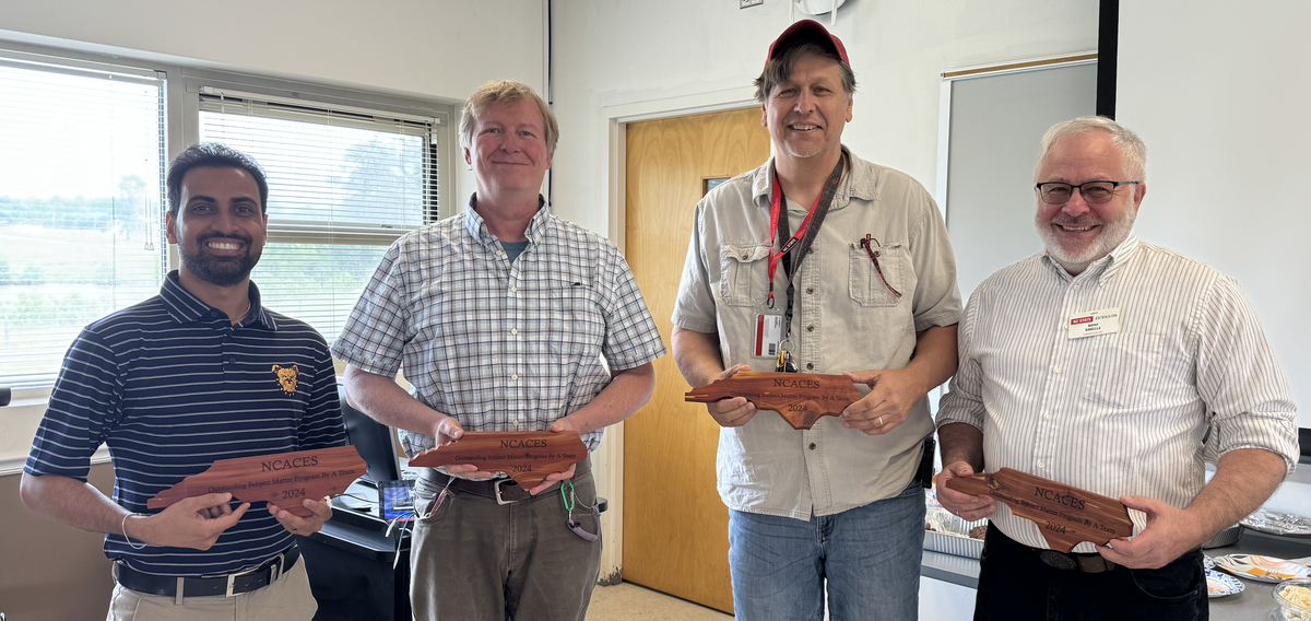 Four men stand next to each other, posing with awards made of wood and shaped like North Carolina. 