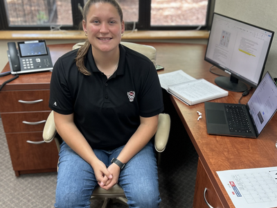 Person sitting in office chair at desk with laptop, monitor, phone, and open notebook.