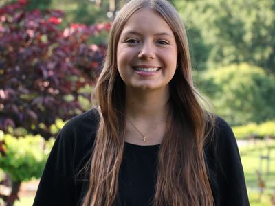 Young woman with long brown hair in black shirt and gold cross necklace, outdoors