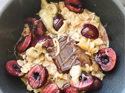 Bowl containing cooked oatmeal, topped with cherries, dark chocolate, peanut butter, and honey.