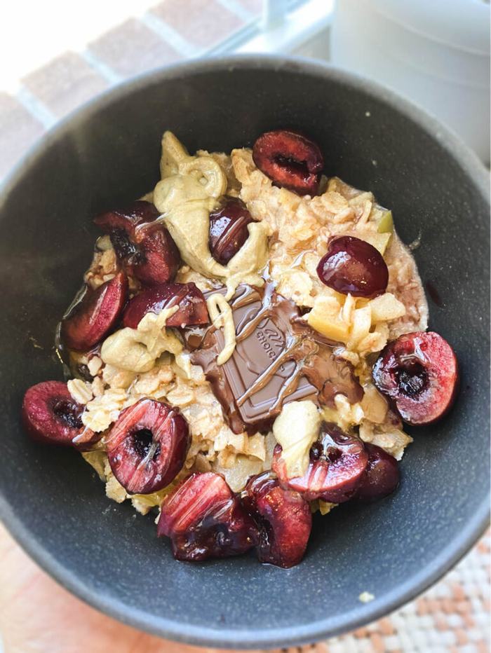 Bowl containing cooked oatmeal, topped with cherries, dark chocolate, peanut butter, and honey. 