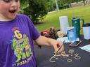 Child at outdoor table using tweezers to thread rubber band and beans; shirt reads "DUDE! I'm 6"