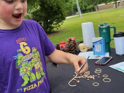 Child at outdoor table using tweezers to thread rubber band and beans; shirt reads "DUDE! I'm 6"