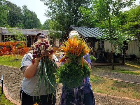 Two people hold large bunches of freshly harvested red onions and carrots covering their faces
