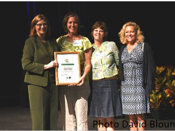 Four women on stage; second from left holds framed certificate; visible text "Photo David Blount"