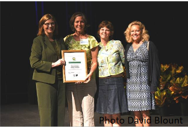 Four women on stage; second from left holds framed certificate; visible text "Photo David Blount"