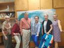 Six adults standing in front of an ocean backdrop, one holding a blue bodyboard.