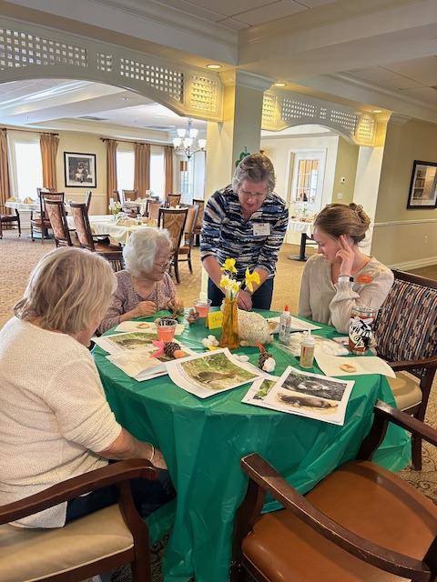 Four women seated around table doing crafts with papers, glue, and daffodils.