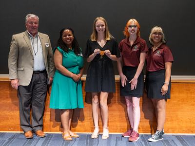 Five people standing in a row, center person holding a medal.