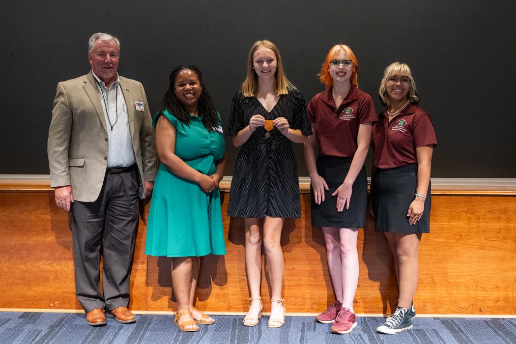 Five people standing in a row, center person holding a medal.