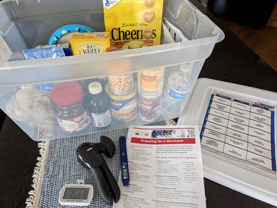 Plastic bin with emergency food (canned goods, Cheerios, water) beside hurricane prep flyer