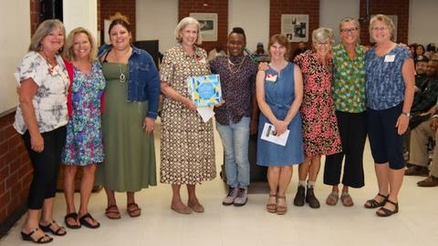 Group of nine people standing indoors, one person holding a blue award plaque