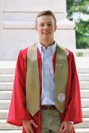 A young man with graduation attire on in front of the NC State Belltower.