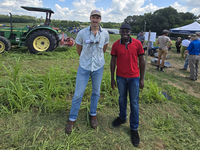 Two men standing in a grassy field near a tractor and people at an outdoor event