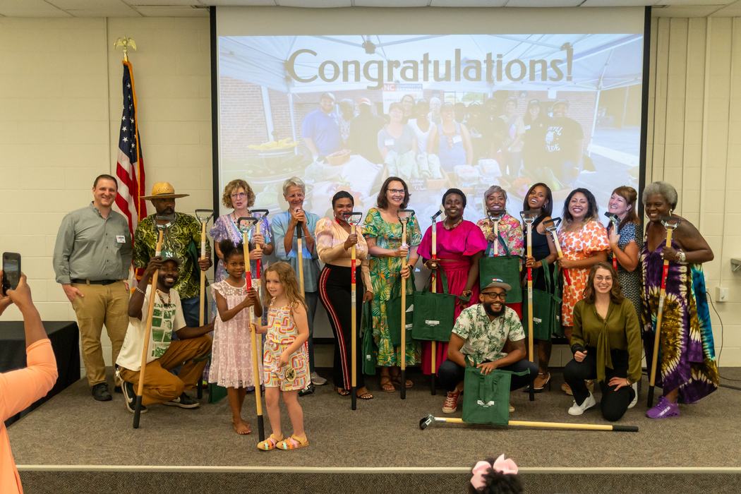 Group of people holding shovels and green bags in front of a screen reading "Congratulations!"