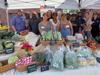 Farmers market stall displaying vegetables and packaged greens with vendors under a tent