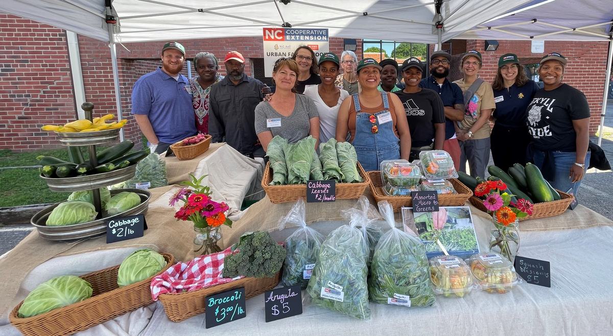 Farmers market stall displaying vegetables and packaged greens with vendors under a tent