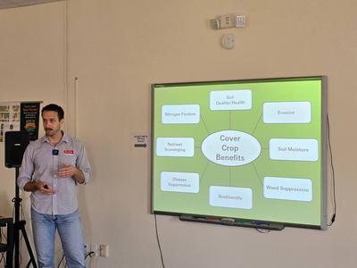 Man presenting slide titled "Cover Crop Benefits" listing soil quality, erosion, moisture, nitrogen.