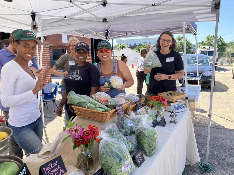 Farmers market vendors under a tent selling packaged leafy greens and vegetables, one holds a bag.