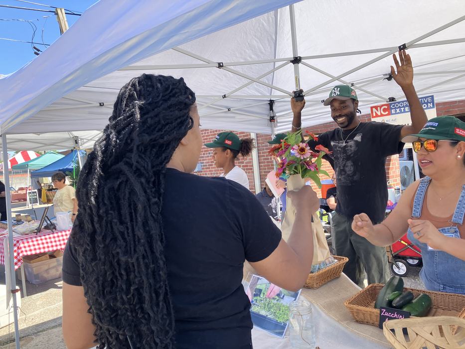 Customer hands bouquet to vendor at farmers market stall under white canopy