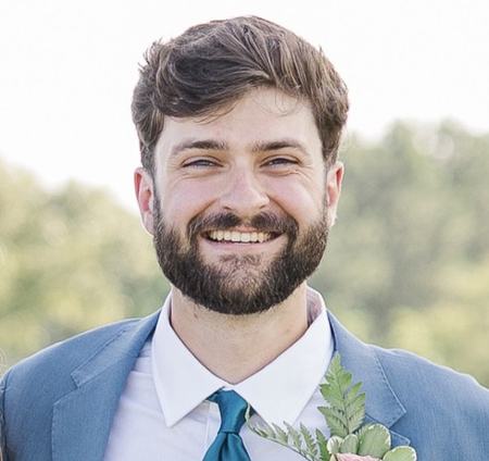 A man smiling with a coat and tie on outdoors on a sunny day.